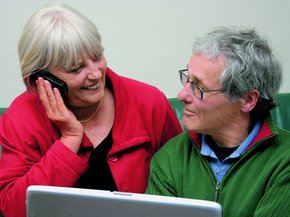 Älteres Paar vor einem Laptop sieht sich an, während die Frau telefoniert